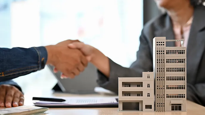 建物模型と書類の前で握手する２人のビジネスシーン。管理会社の変更や管理プラン・費用の相談をイメージさせる写真。