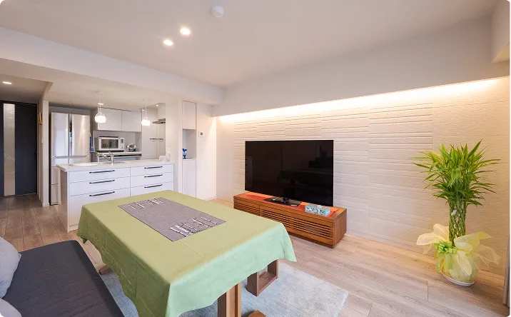 マンションのリフォーム事例。モダンなリビング・ダイニング・キッチンで、白い壁に大型テレビ、緑のテーブルクロスをかけたダイニングテーブル、観葉植物、白いキャビネットとステンレス家電が並ぶオープンキッチン。