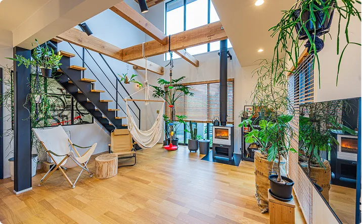 一戸建てのリフォーム事例。高天井と木製梁が特徴のモダンなリビング空間で、黒い金属階段、ハンモックチェア、暖炉、観葉植物が配置され、自然光が差し込む開放的なインテリア。