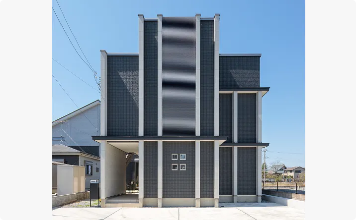 注文住宅の事例。直線的で幾何学的なデザインが特徴のモダンな一戸建て住宅で、中央に縦長パネル、対称的な柱、小窓が並ぶ外観。青空と周囲の住宅街が背景に広がる。